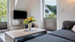 Una sala moderna con un televisor de pantalla plana, un florero con flores, un sofá y una ventana con vista a la vegetación.