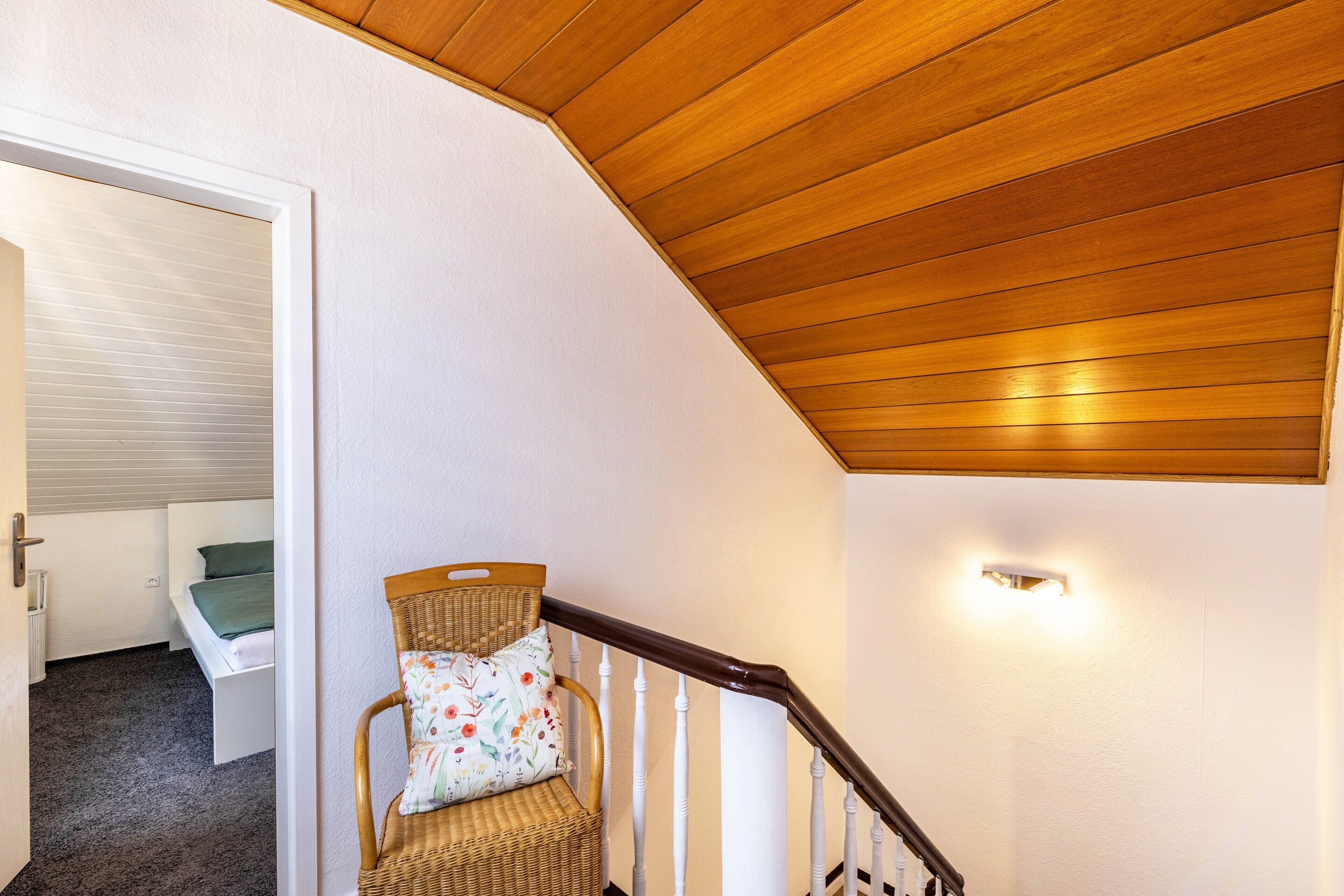 A staircase with a wooden handrail, a wicker chair with a floral cushion, and a bed with a green blanket in the background.