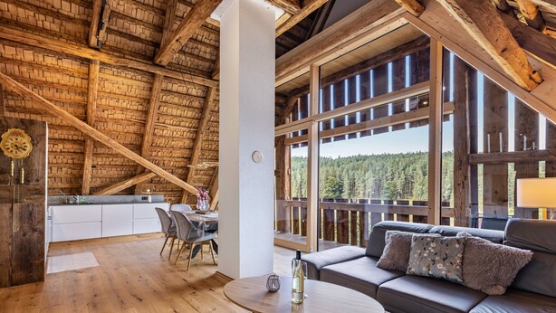 A wooden interior with a dining area, a sofa with cushions, and a large window offering a forest view.