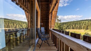 A wooden deck with chairs and a glass balustrade overlooking a forested landscape.