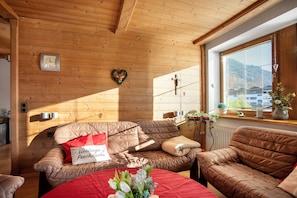 A cozy room with wooden walls, a brown leather sofa, a red table, and a window with blinds.