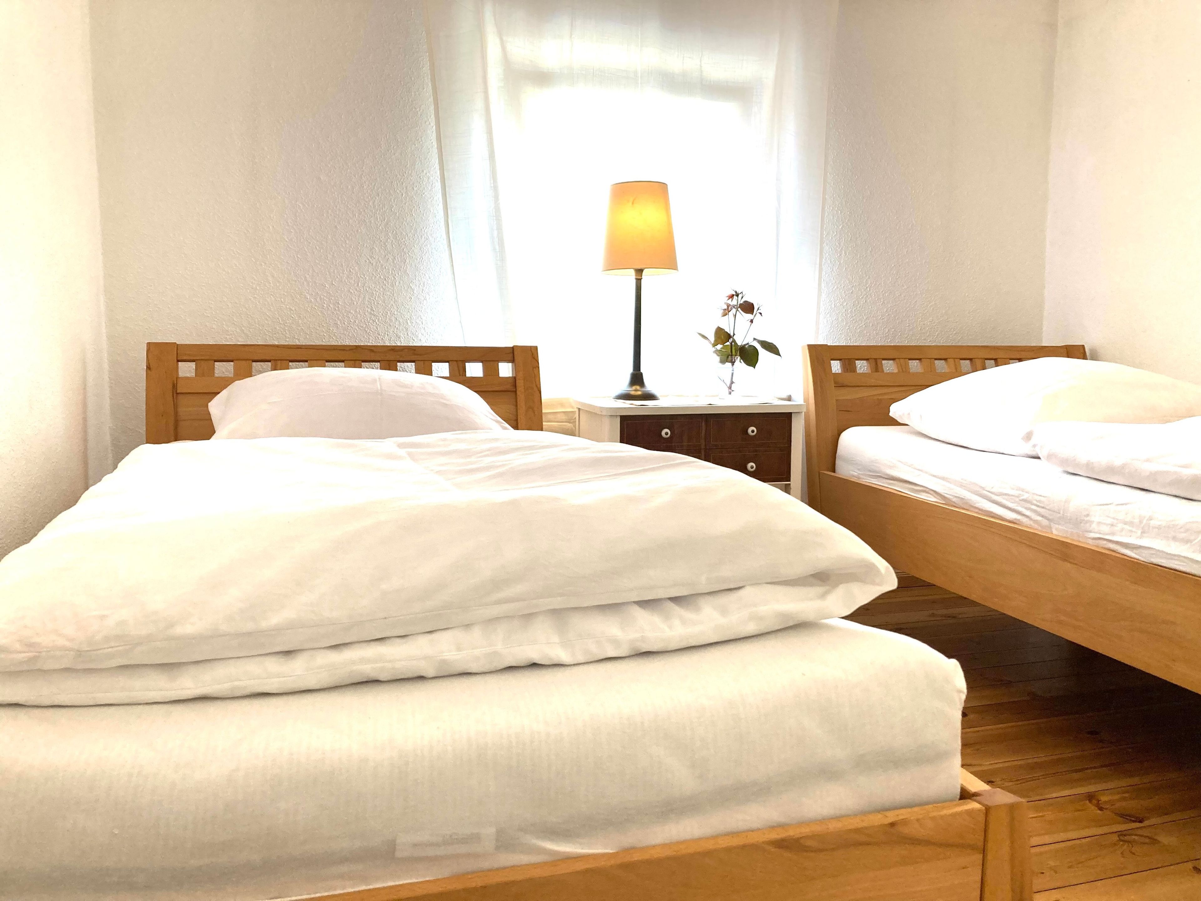 Ein Zimmer mit zwei hölzernen Betten, weißer Bettwäsche, einem Nachttisch mit Lampe und einem Fenster mit durchscheinenden Vorhängen.