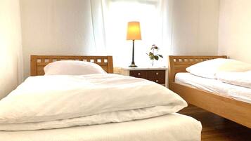 Ein Zimmer mit zwei hölzernen Betten, weißer Bettwäsche, einem Nachttisch mit Lampe und einem Fenster mit durchscheinenden Vorhängen.