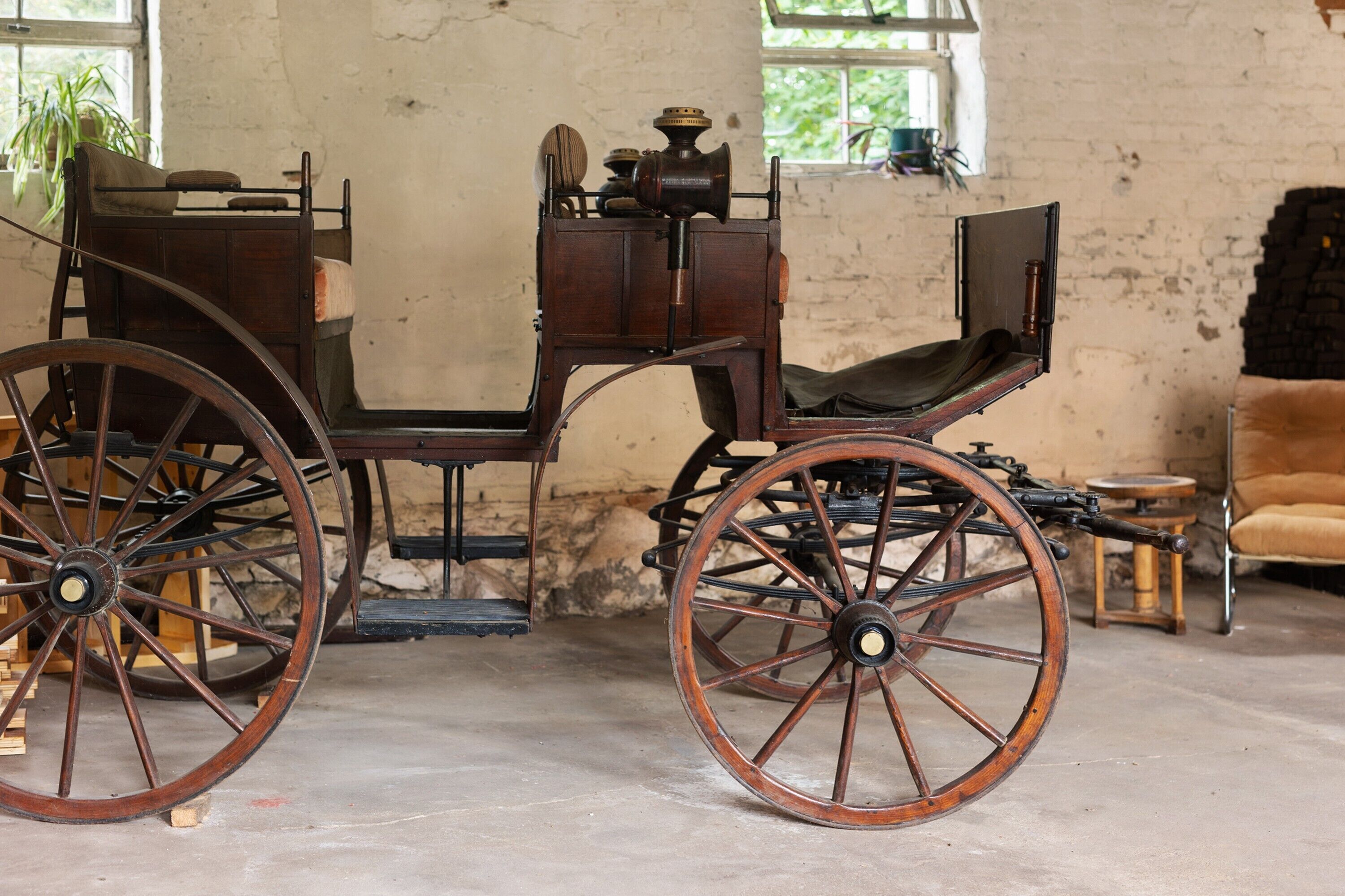 Ein historischer Wagen mit großen Holzwielen und einer Sitzbank, präsentiert in einem Innenraum mit Backsteinwand und Fenster im Hintergrund.