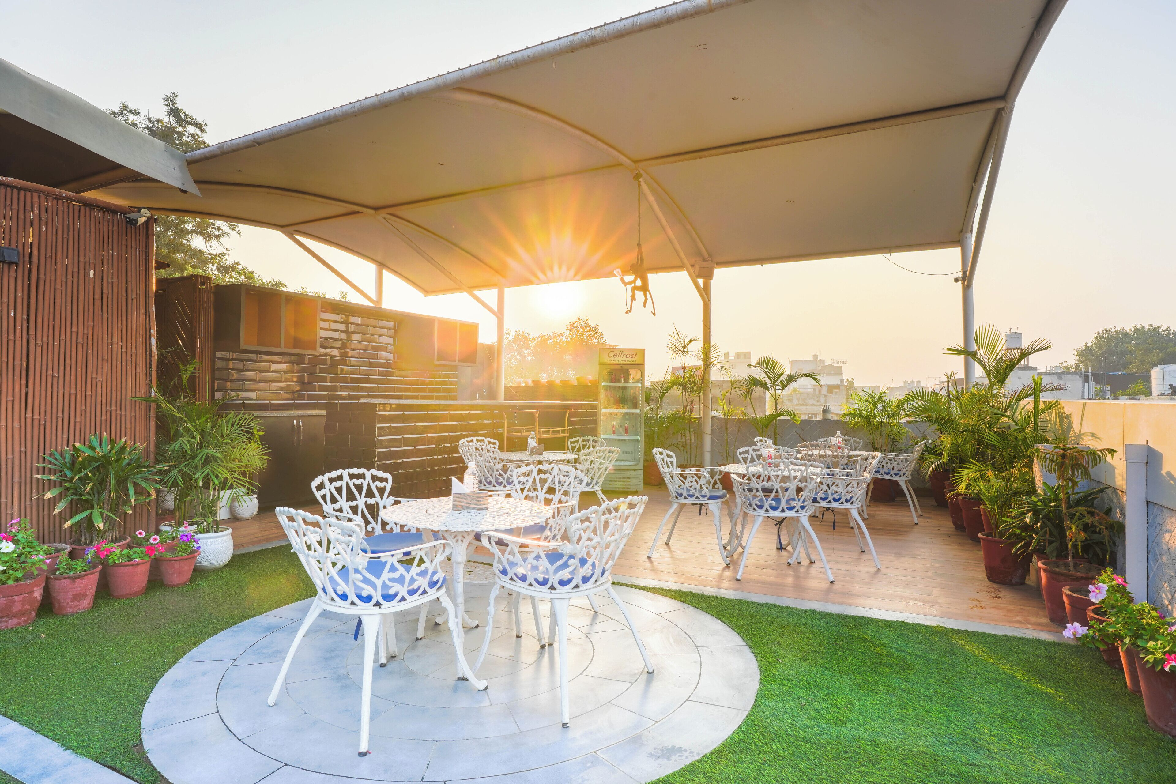 Une terrasse avec des chaises et des tables en métal blanc, entourée de plantes en pot et d’une pelouse.