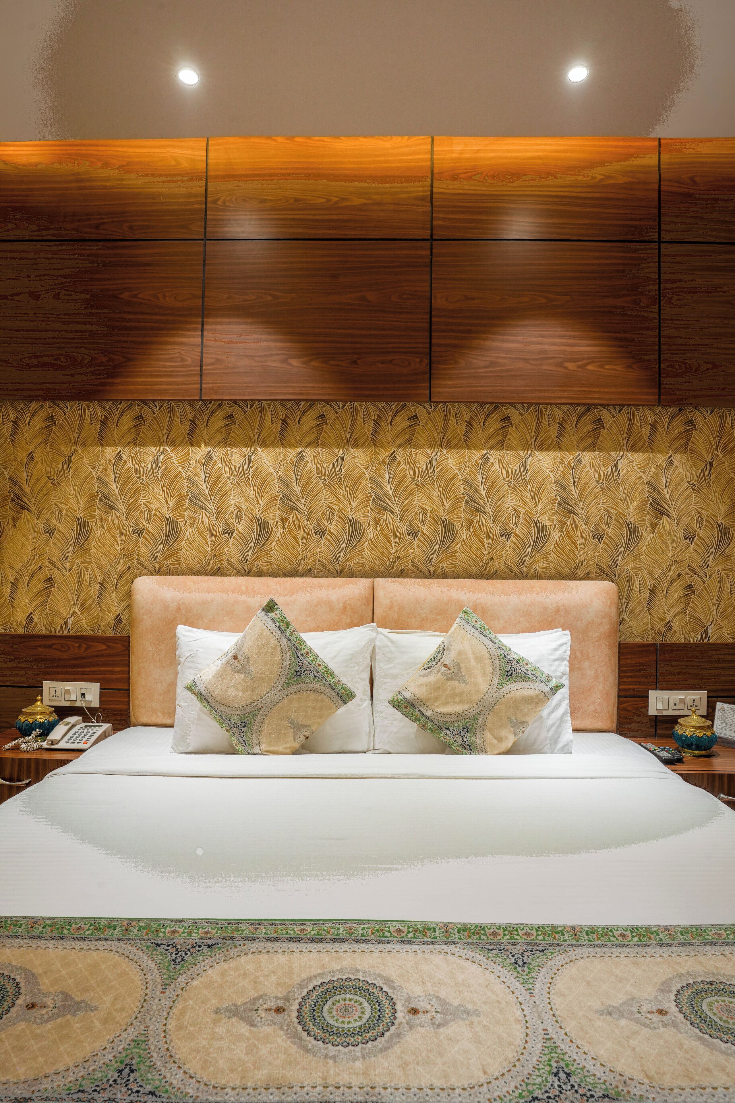 A bedroom with a wooden headboard, two decorative pillows, a patterned rug, and a nightstand with a lamp and a telephone.