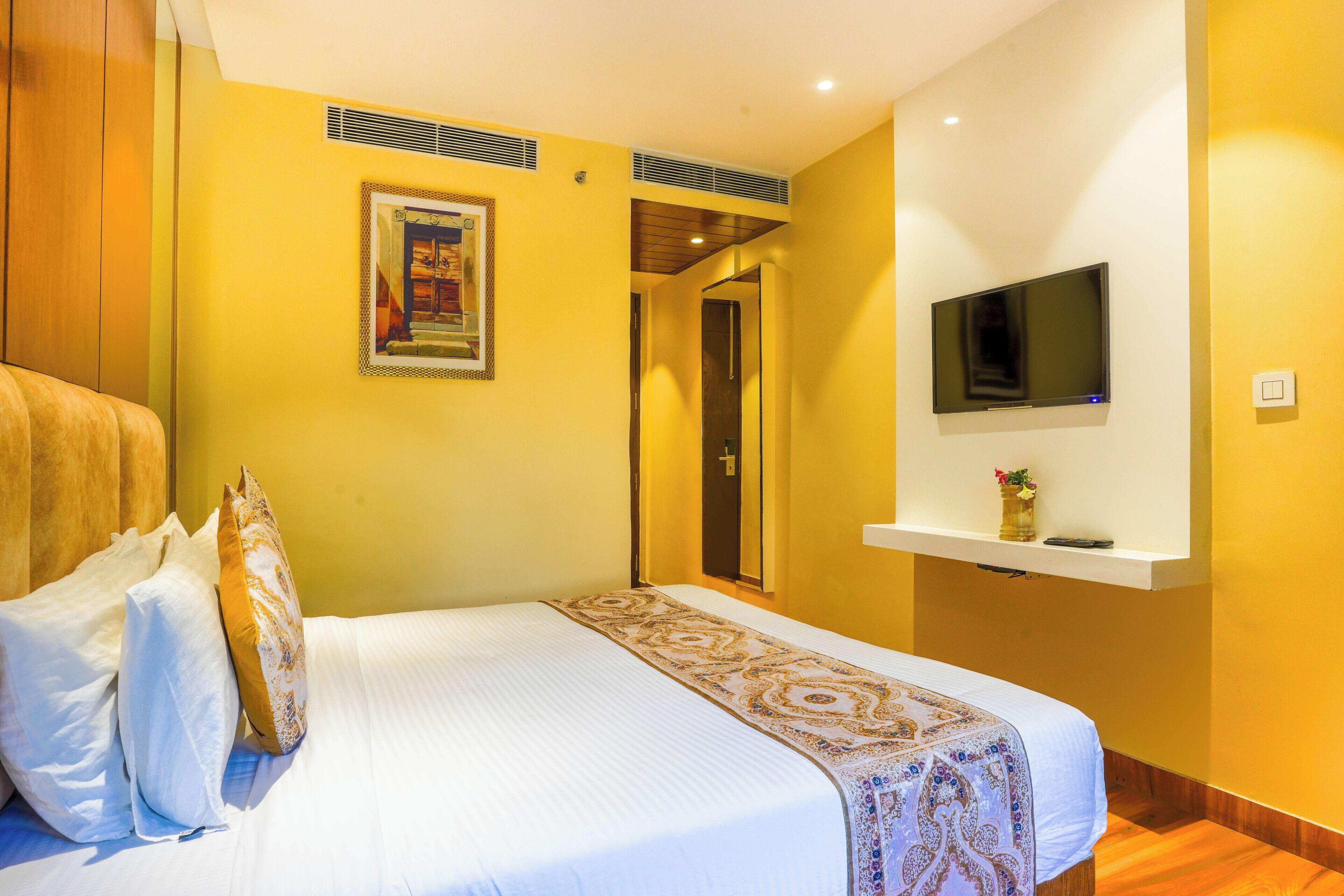 Habitación de hotel con una cama, almohadas, un televisor montado y un cuadro en la pared.