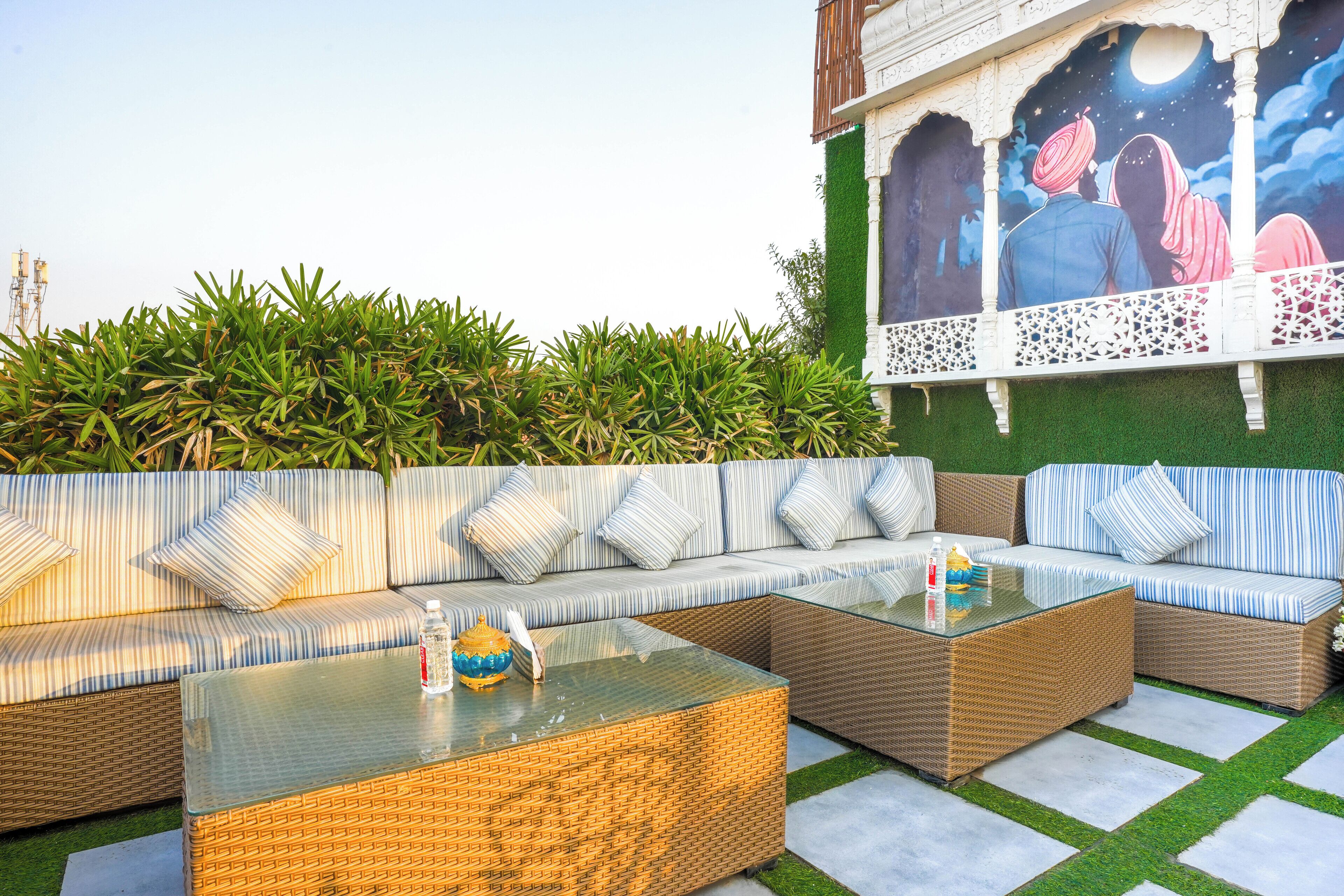 A section of a hotel with a large outdoor seating area featuring wicker sofas and glass-top tables, a mural on the wall, and a lush green hedge in the background.