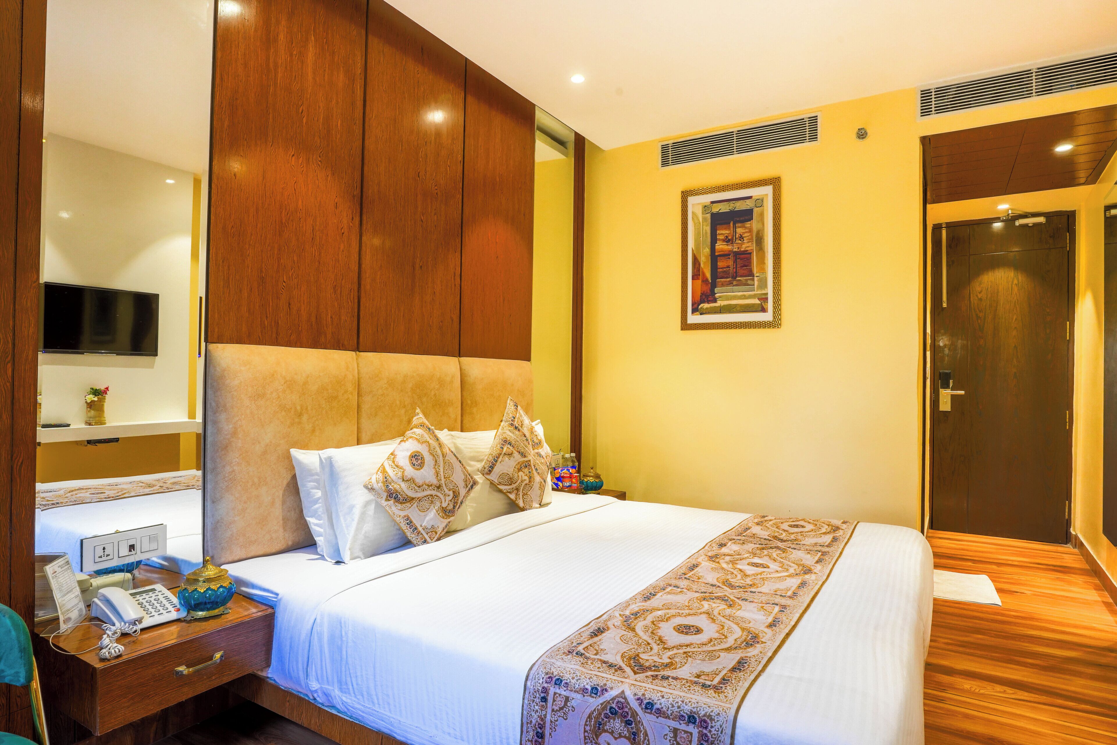 Une chambre d’hôtel avec un grand lit, une tête de lit en bois, une table de chevet avec un téléphone et une lampe, un téléviseur fixé au mur et une porte avec un miroir.