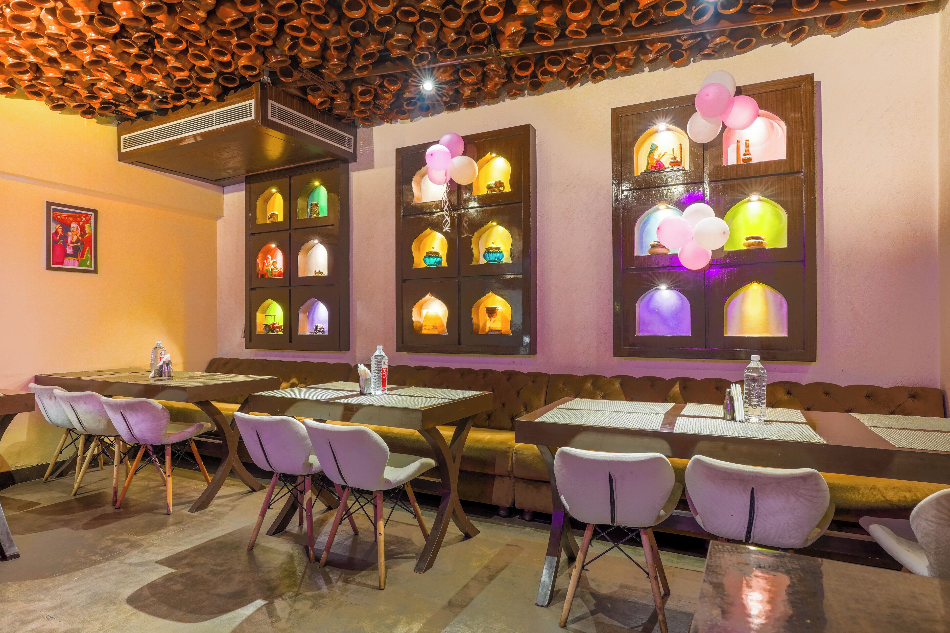 Un restaurante con varias mesas y sillas, un techo con numerosas macetas colgantes y ventanas iluminadas con luces y globos de colores.