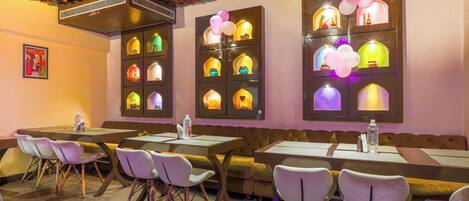 Un restaurante con varias mesas y sillas, un techo con numerosas macetas colgantes y ventanas iluminadas con luces y globos de colores.