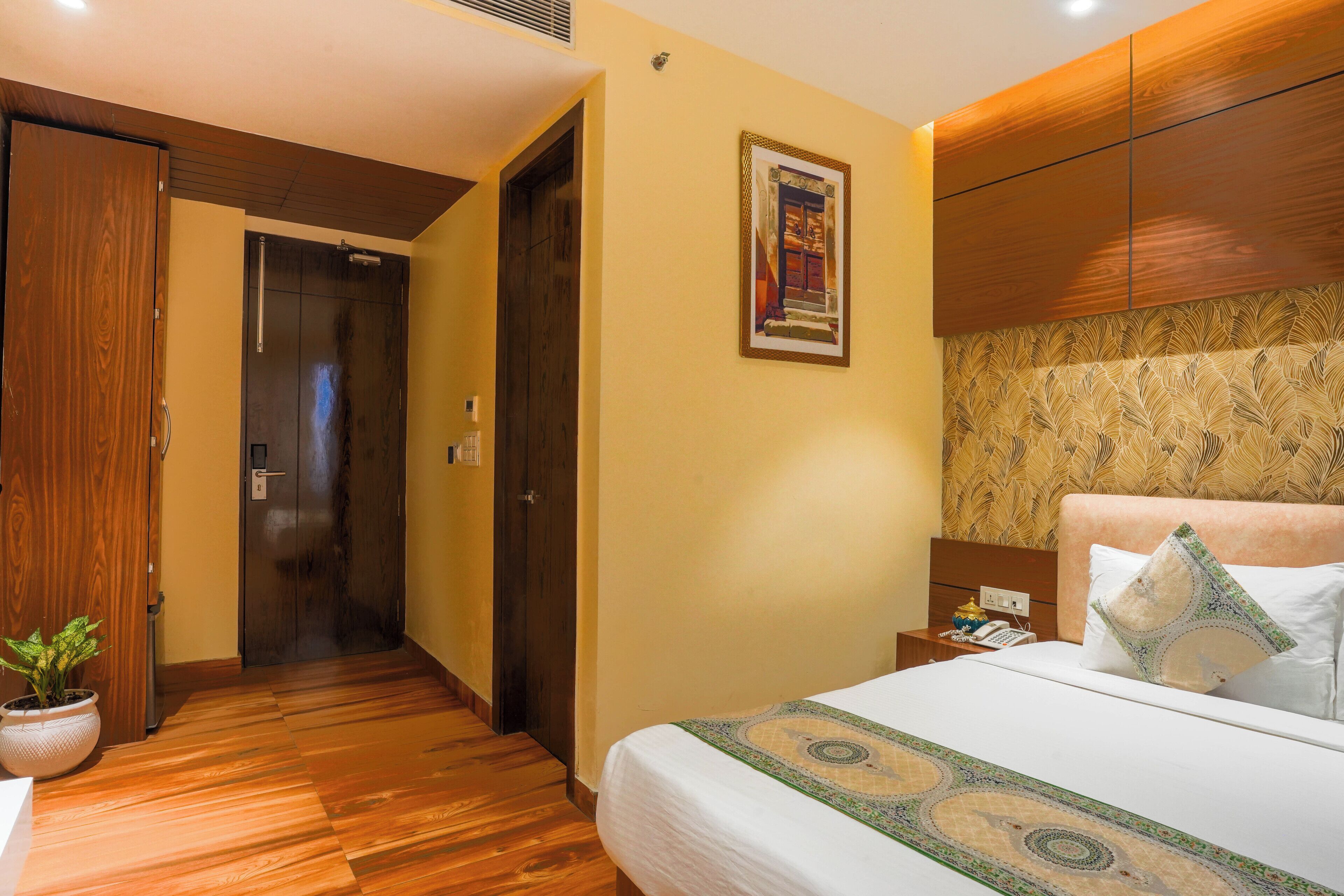 A hotel room with a wooden floor, a bed with a patterned bedspread, a nightstand with a phone, and a wall-mounted cabinet.