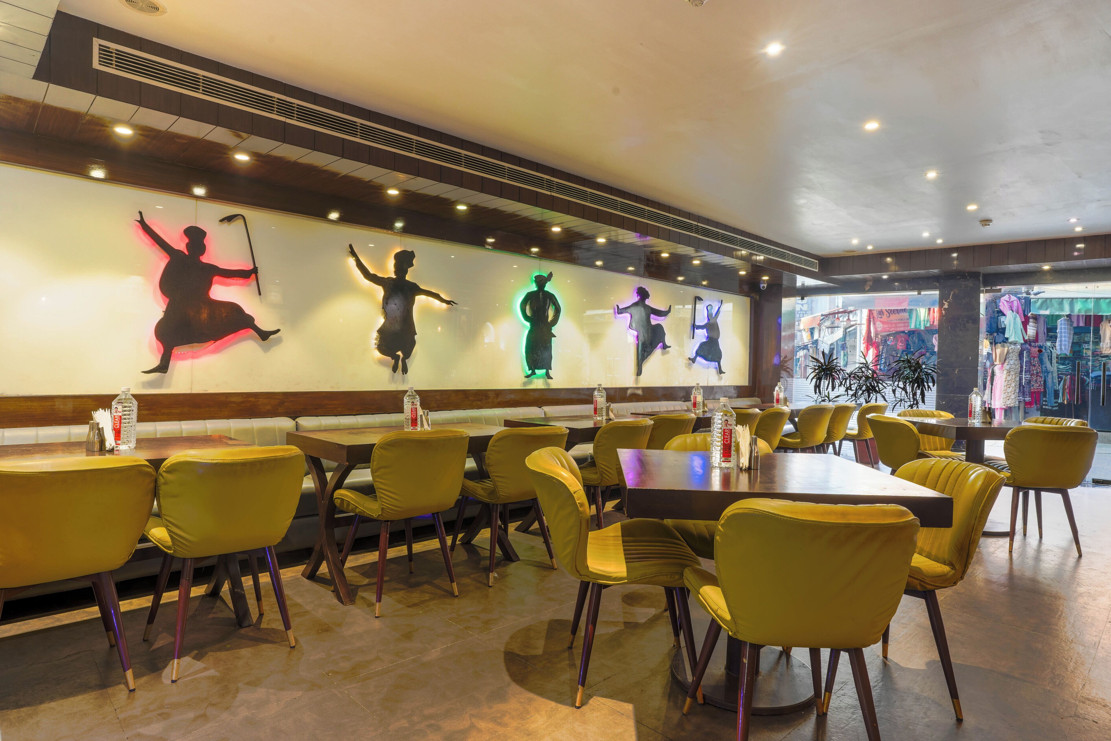 A restaurant with yellow chairs and tables, a wall with illuminated silhouettes of dancers, and a view of the street outside.