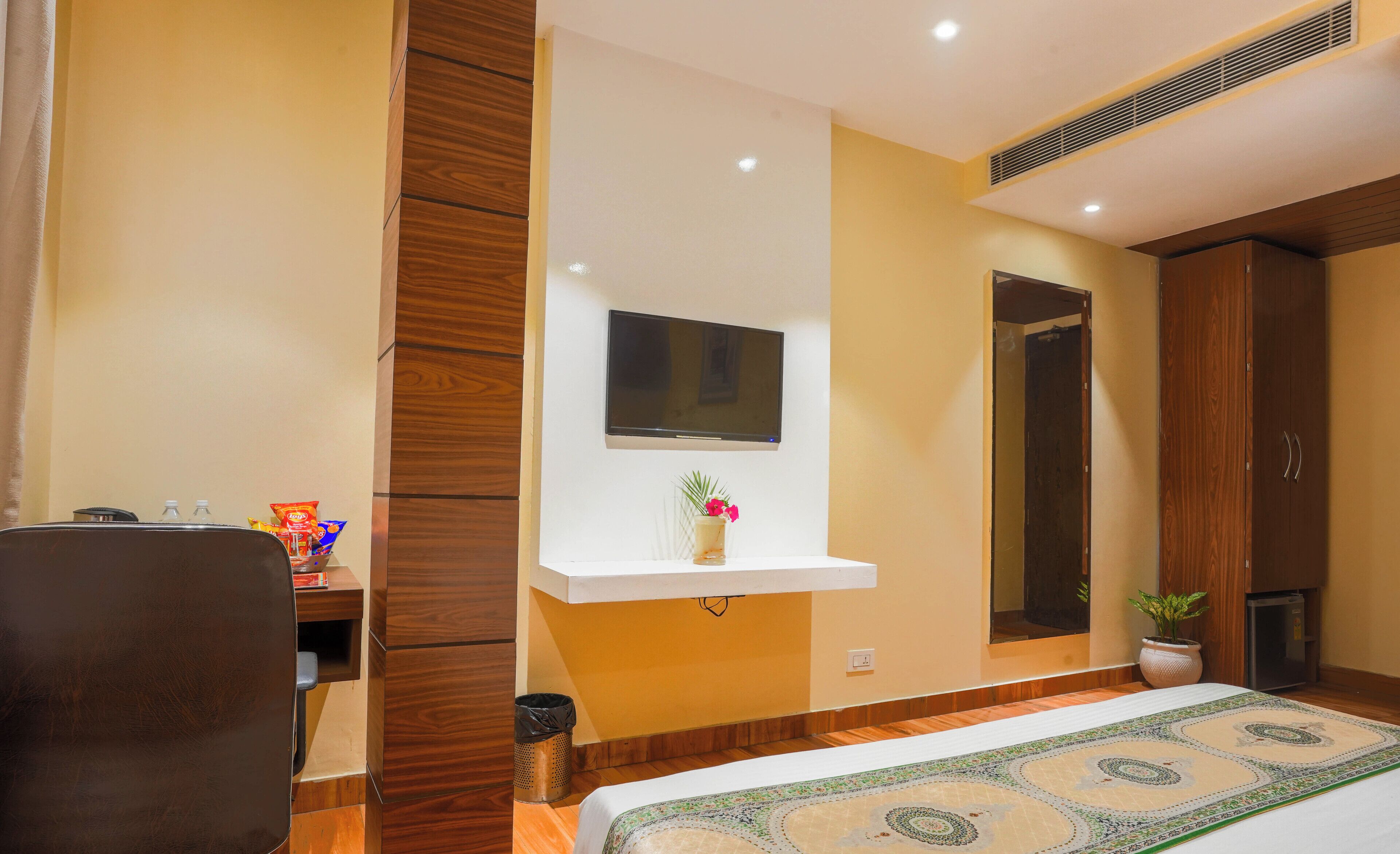 A hotel room with a bed, a desk, a TV mounted on the wall, and a potted plant.