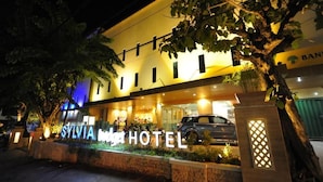 Een hotel genaamd “SYLVIA HOTEL” met een moderne gevel, verlichte reclameborden en een geparkeerde auto ervoor.