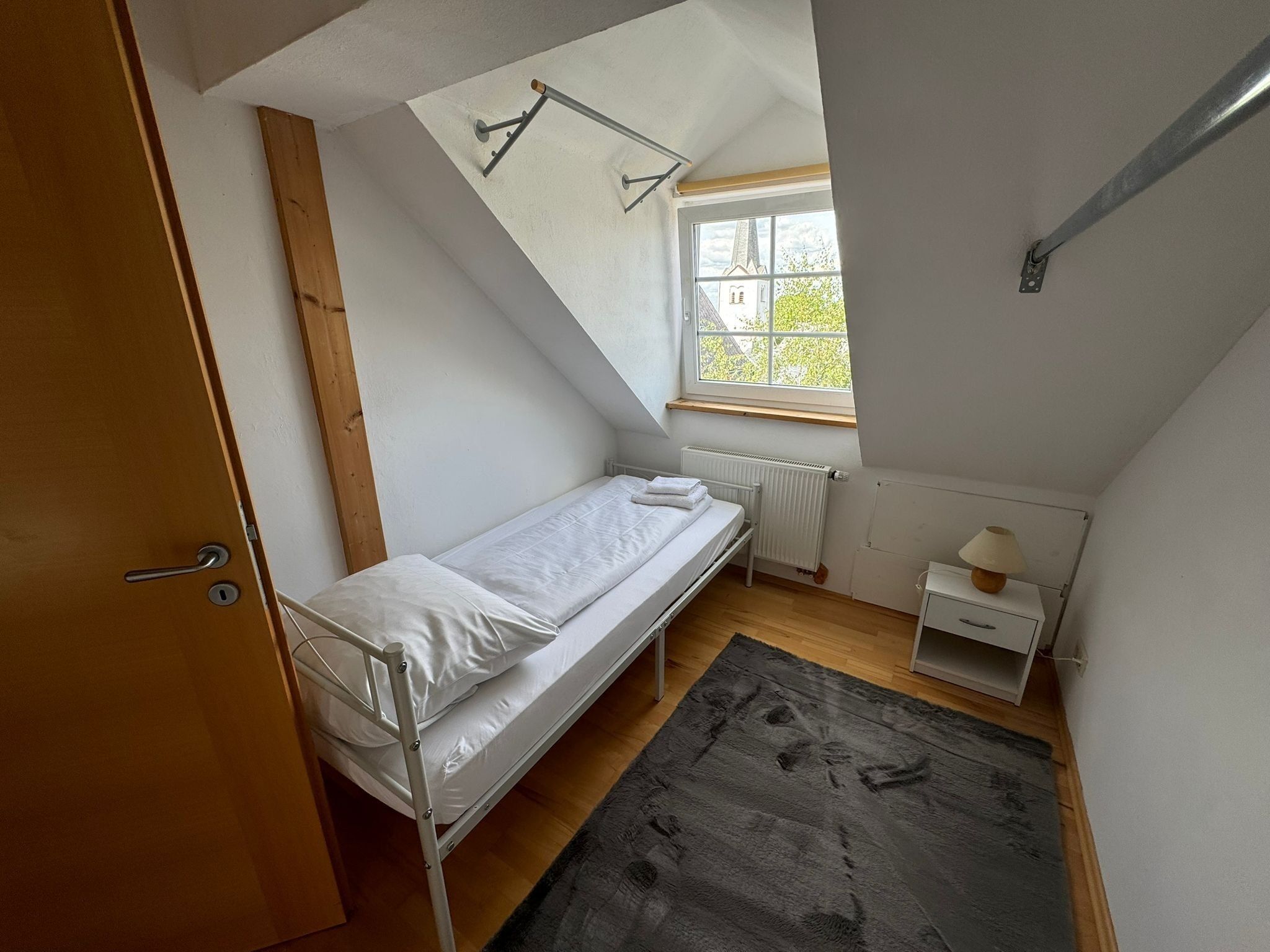 Pequeña habitación individual con techo inclinado, una ventana, un radiador, una mesita de noche con lámpara y una puerta.