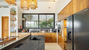 A modern kitchen with a large island, stainless steel appliances, wooden cabinets, and a large window offering a view of greenery.