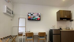 A compact kitchen with a white countertop, two wooden chairs, a small refrigerator, and a wall-mounted TV displaying various channels. - Melce Place Studio Unit Condo (Lapu-Lapu City)