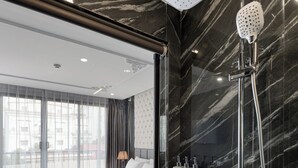 Ein modernes Hotelzimmer mit einem großen Bett, einer Dusche inklusive Handbrause und Blick auf die Stadt durch große Fenster.