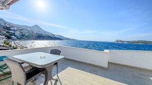 Une terrasse avec une table et des chaises donnant sur une zone côtière, avec des bâtiments et une montagne en arrière-plan.