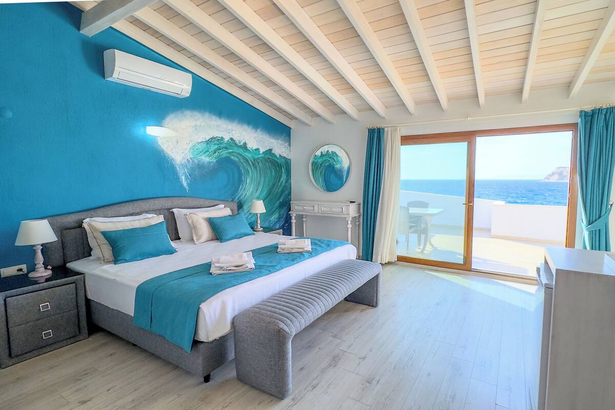 A bedroom with a bed, bedside tables, a desk, and a view of the sea through a glass door.