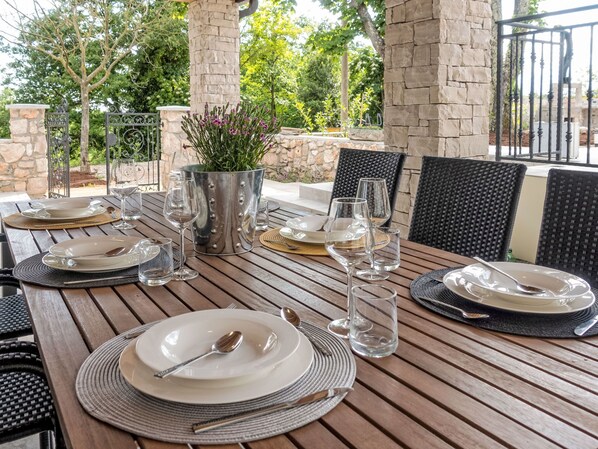 An outdoor dining area with a stone pillar, metal railing, and a table set for four with plates, glasses, and cutlery.