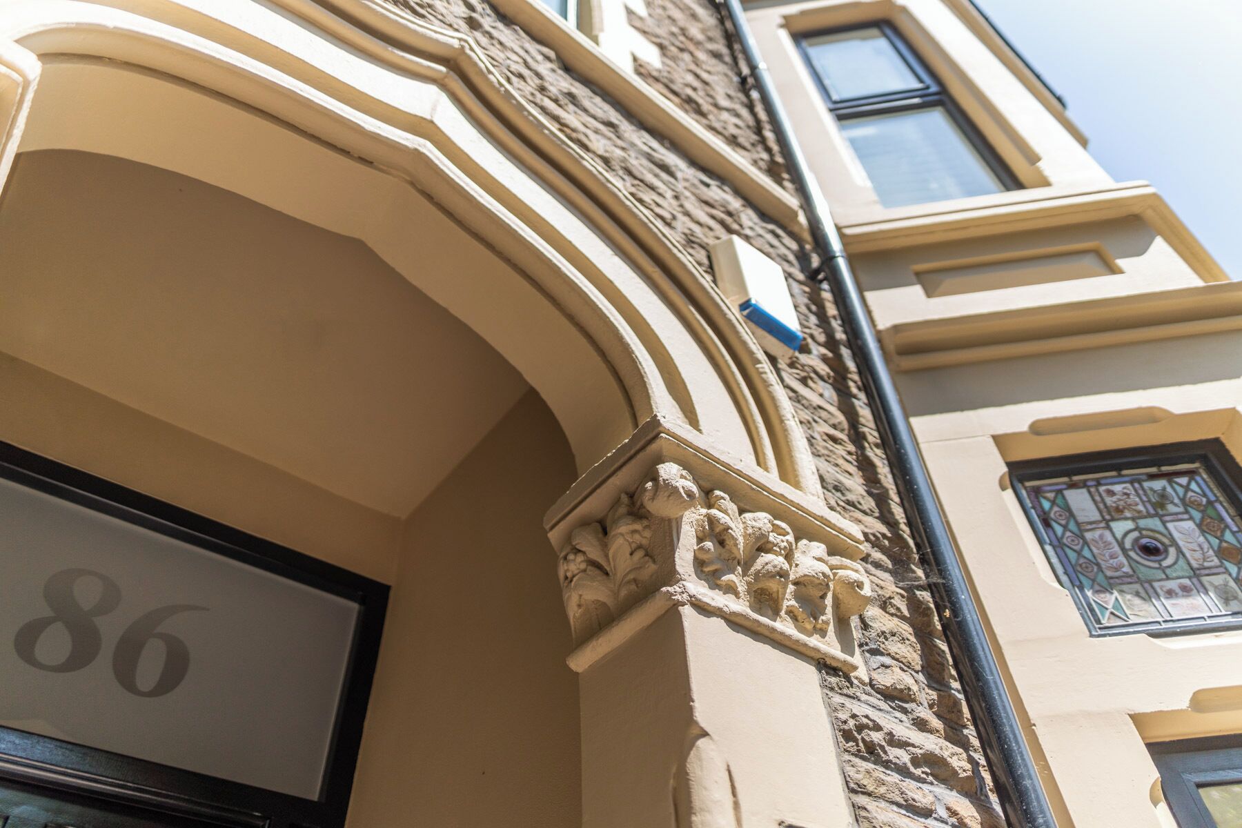 Esquina de un edificio con fachada de piedra, una ventana con vidrieras y una puerta con un número.