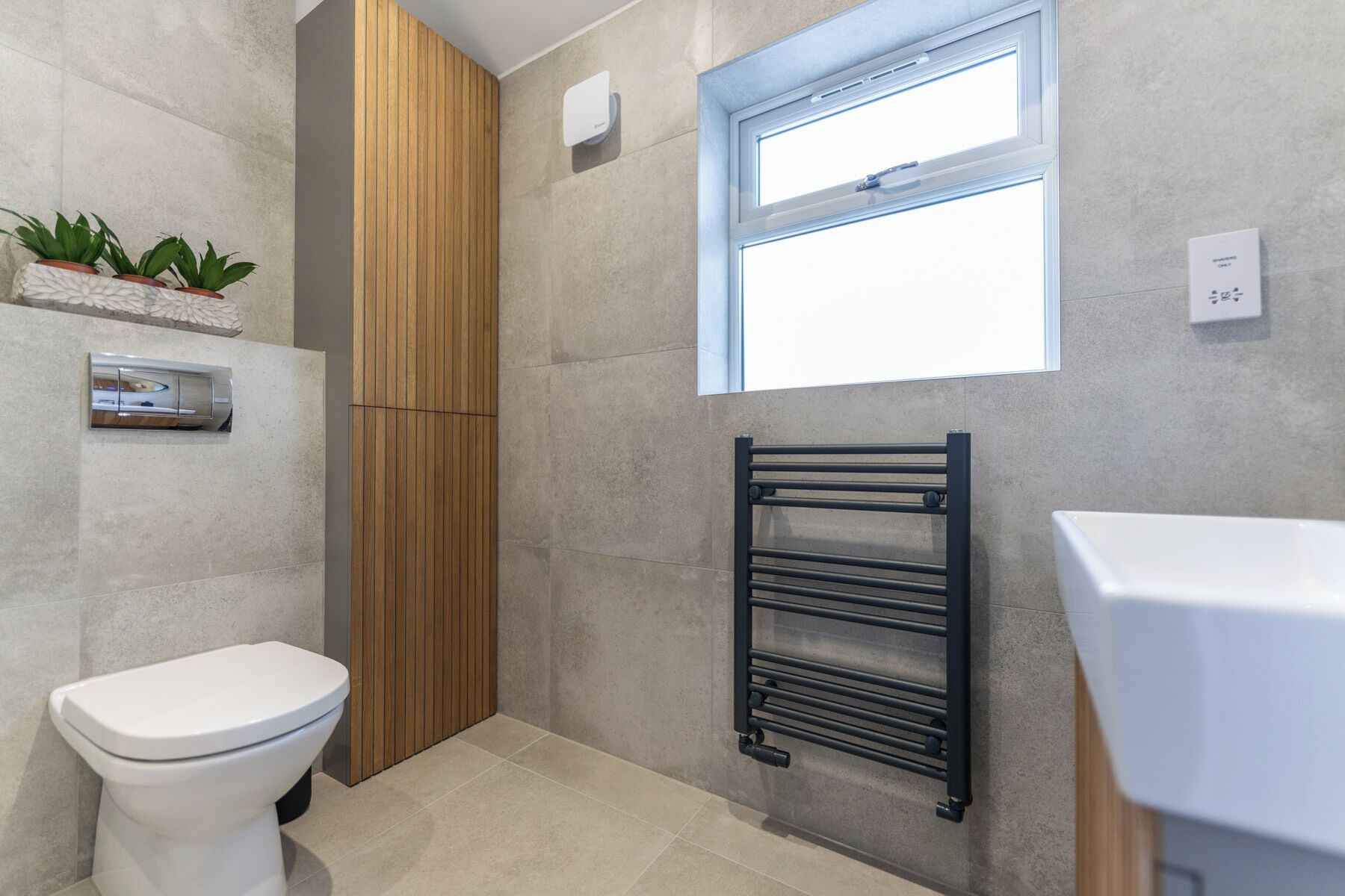 Un baño moderno con inodoro, pared de paneles de madera, toallero negro, ventana y estante pequeño con plantas.