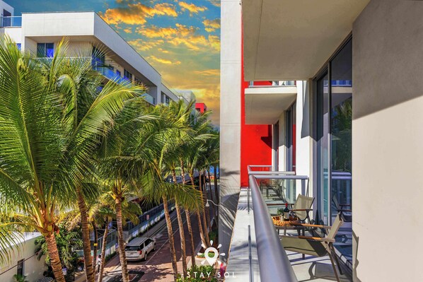 Un hotel moderno con fachada roja y blanca, un balcón con mobiliario y palmeras al frente.