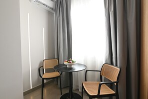 A room with two wicker chairs, a small round table with a bowl of fruit, and a window with grey curtains. - Deos living experience 1 (Athina)
