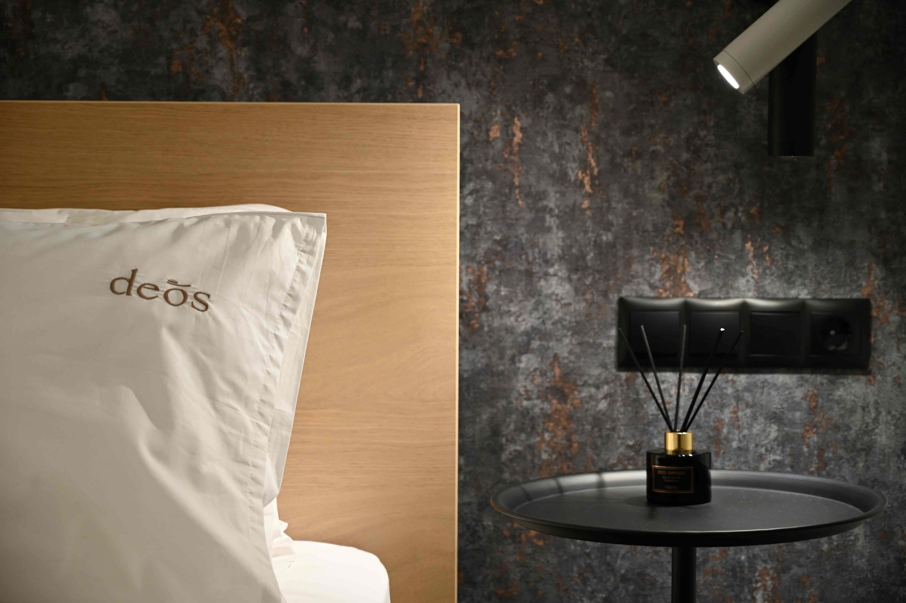 Quarto de hotel com cama, travesseiro branco com o texto 'deos', mesa redonda com difusor preto e parede texturizada.