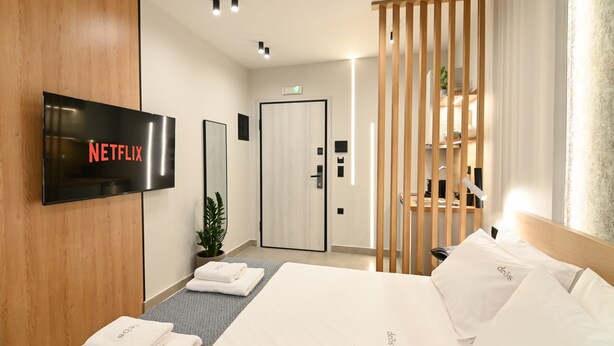 A hotel room with a bed, a TV mounted on the wall, a wooden partition, and a door leading to a bathroom. - Deos living experience 1 (Athina)