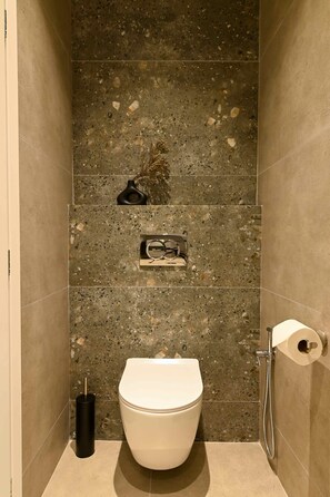 A bathroom with a toilet, a shower, and a small shelf with a decorative item. - Deos living experience 1 (Athina)