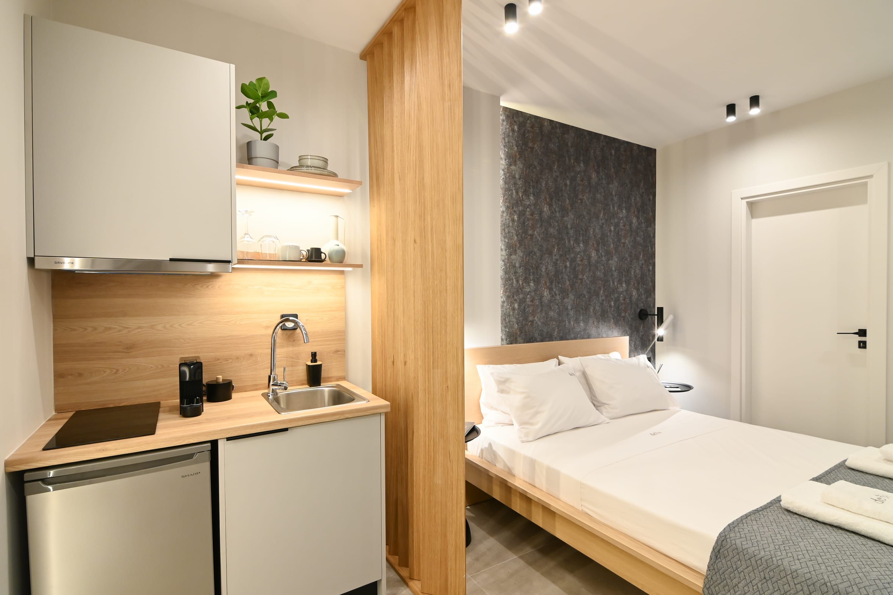 Een compacte hotelkamer met kitchenette, een bed met witte beddengoed en een donkere, gestructureerde wand.