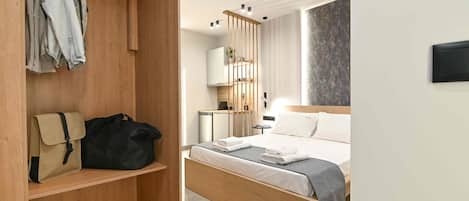 A hotel room with a wooden bed, a wardrobe with hanging clothes, a small desk, and a bedside table.