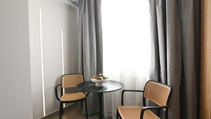 A small room with a round table and two wicker chairs, a bowl of fruit on the table, and a window with grey curtains. - Deos living experience 1 (Athina)