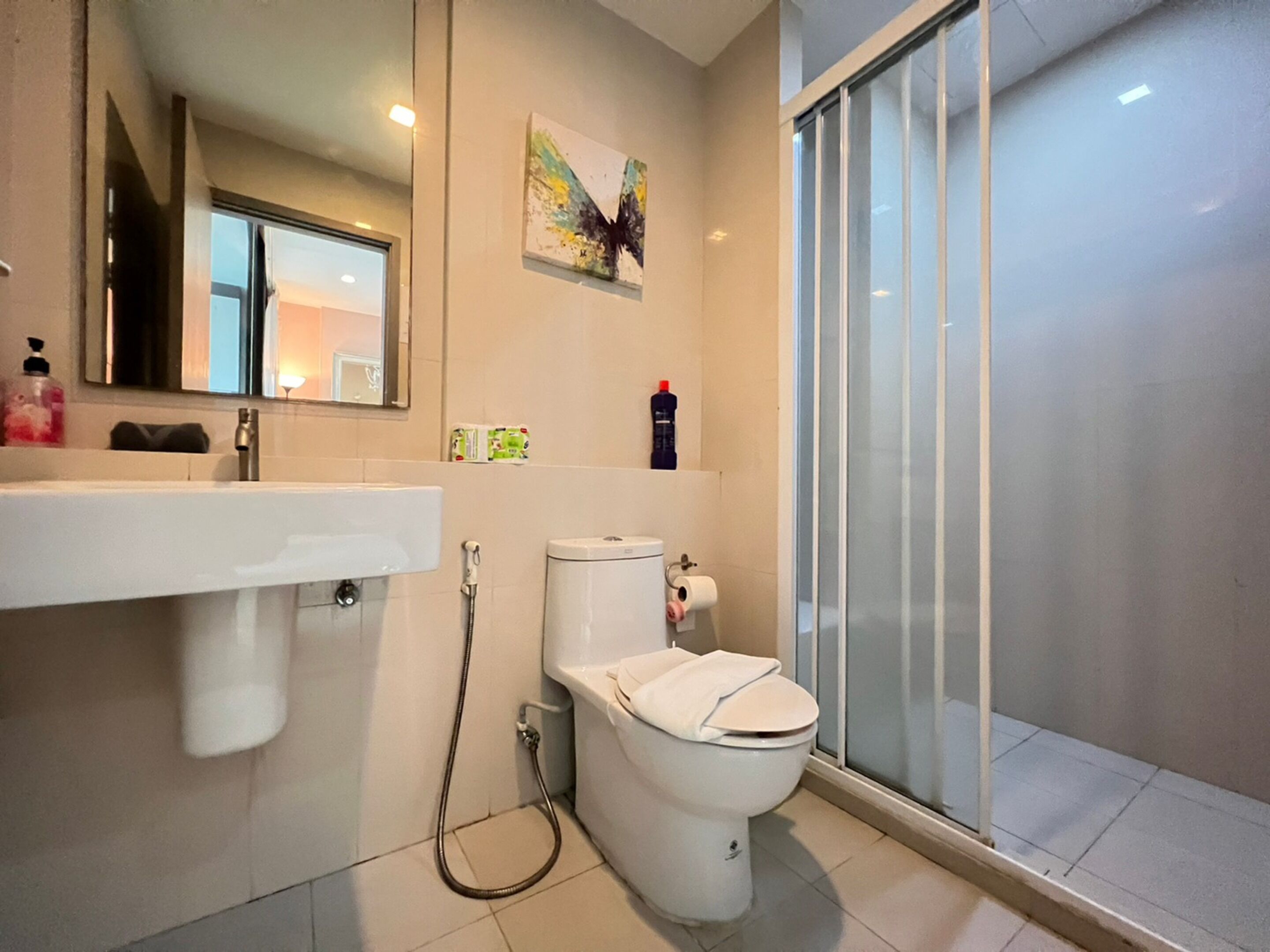 A modern bathroom with a white sink, toilet, and shower. There is a mirror above the sink and a painting on the wall.