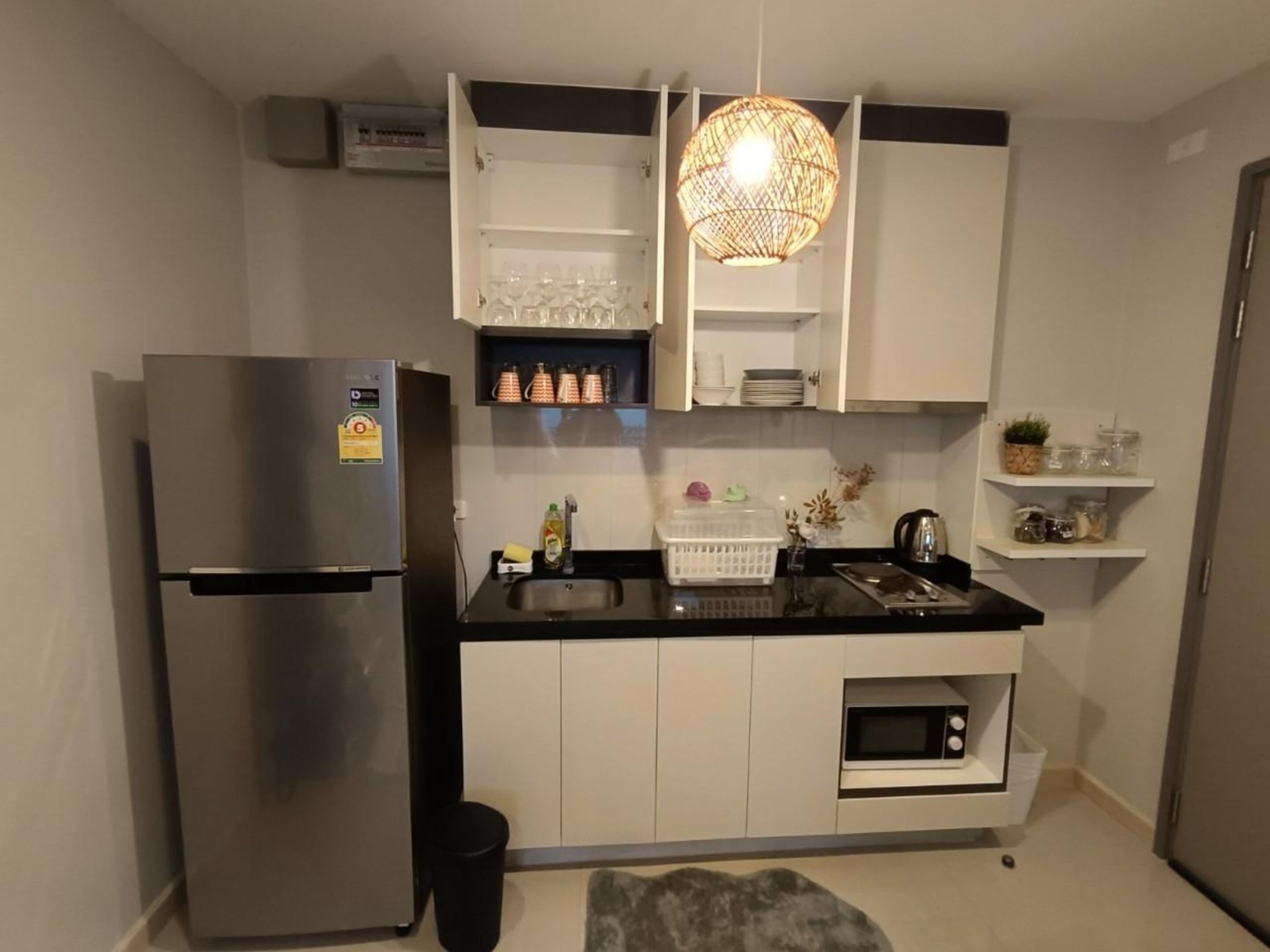 A compact kitchen with a stainless steel refrigerator, a microwave, and a built-in oven. There are white cabinets, a black countertop, and a hanging light fixture.