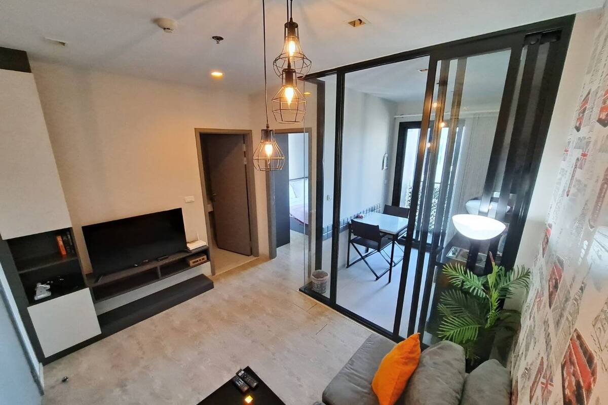 A modern living room with a flat-screen TV, a sofa with an orange cushion, a coffee table, and a dining area with a table and chairs.
