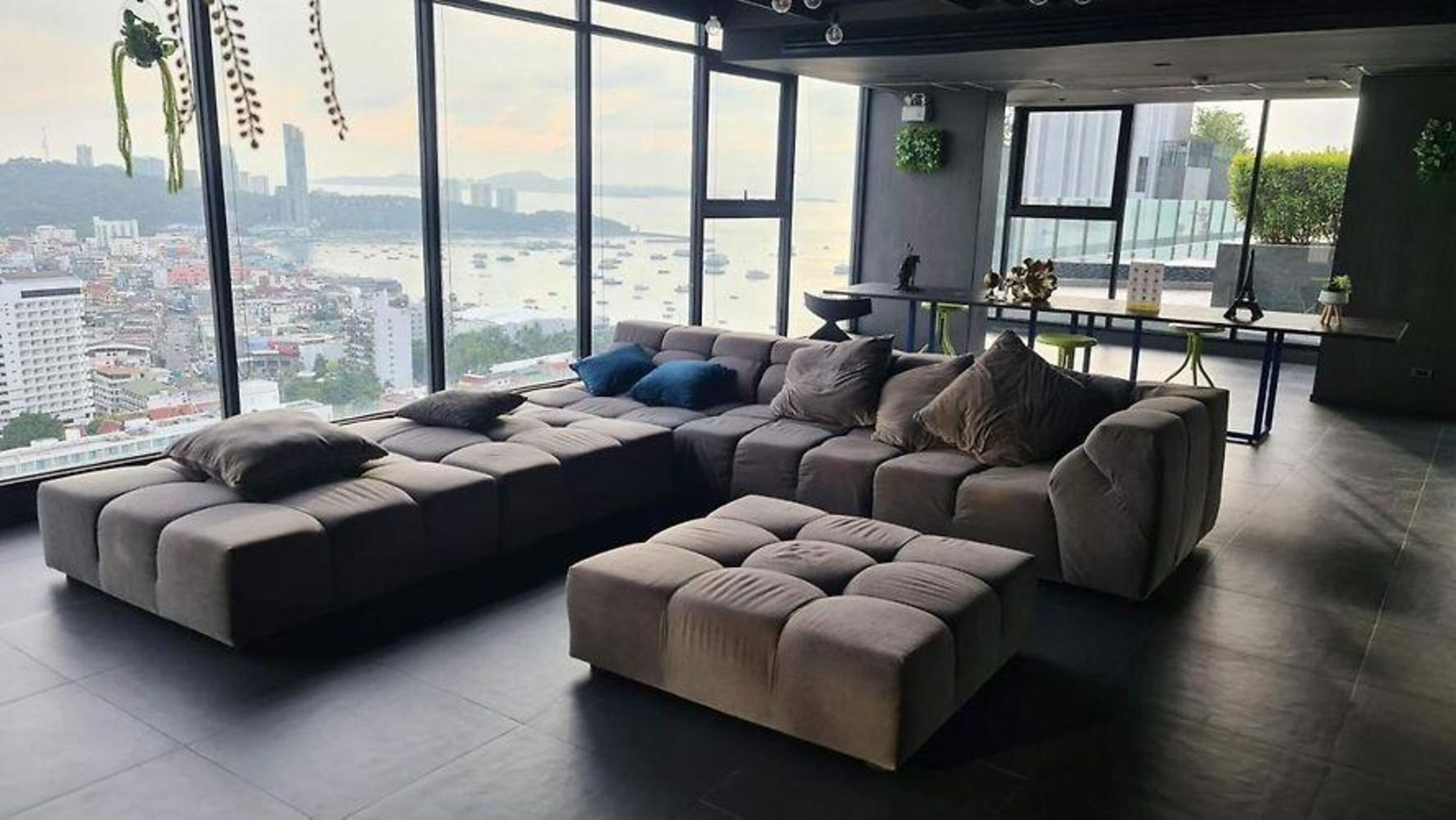 Ein modernes Wohnzimmer mit Ecksofa und Hocker, großen Fenstern mit Blick auf die Stadt und einem kleinen Beistelltisch mit Pflanze.