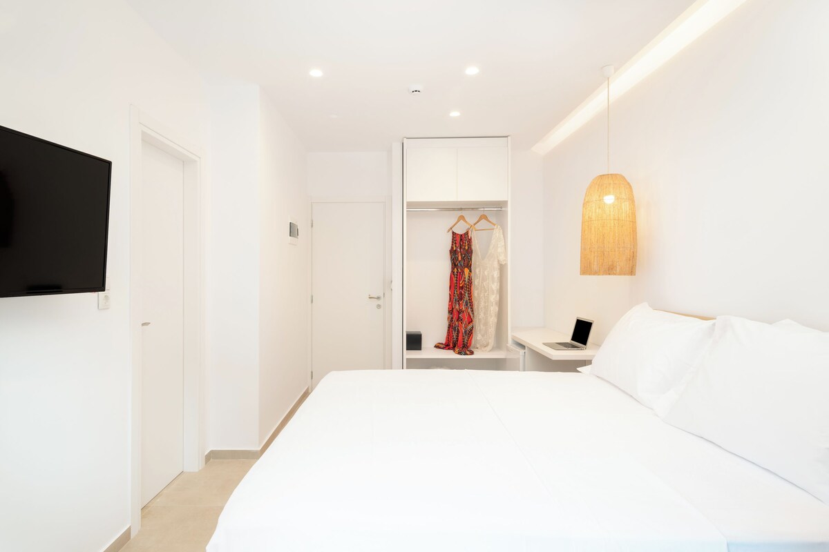 A modern hotel room with a large bed, a TV mounted on the wall, a wardrobe, and a bedside table with a lamp.