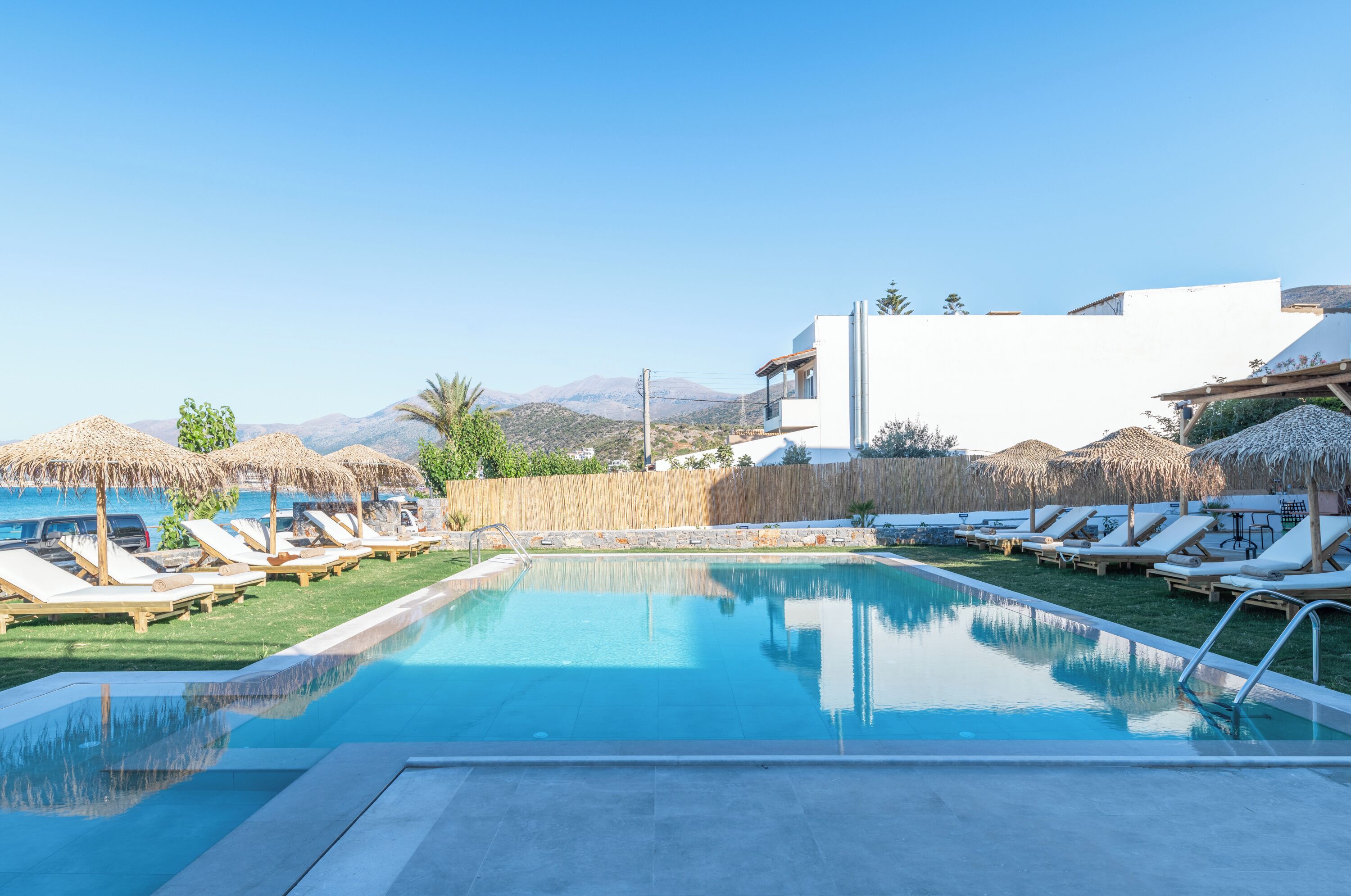 Una piscina con sillas de descanso y sombrillas, rodeada por una valla de madera y un edificio blanco.