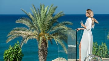 Una mujer con un vestido blanco de pie junto a una barandilla de cristal con vistas a una piscina, palmeras y el mar al fondo.