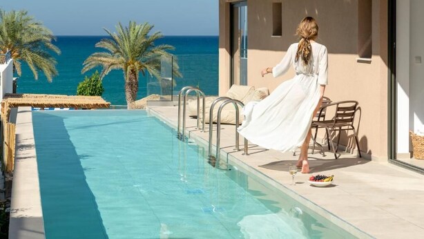 Zona junto a la piscina con vista al mar, una mujer con un vestido blanco y mobiliario de exterior.