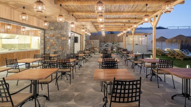 An outdoor dining area with wooden tables and chairs, stone walls, and hanging lights. - Golden Bee Lifestyle (Hersonissos)