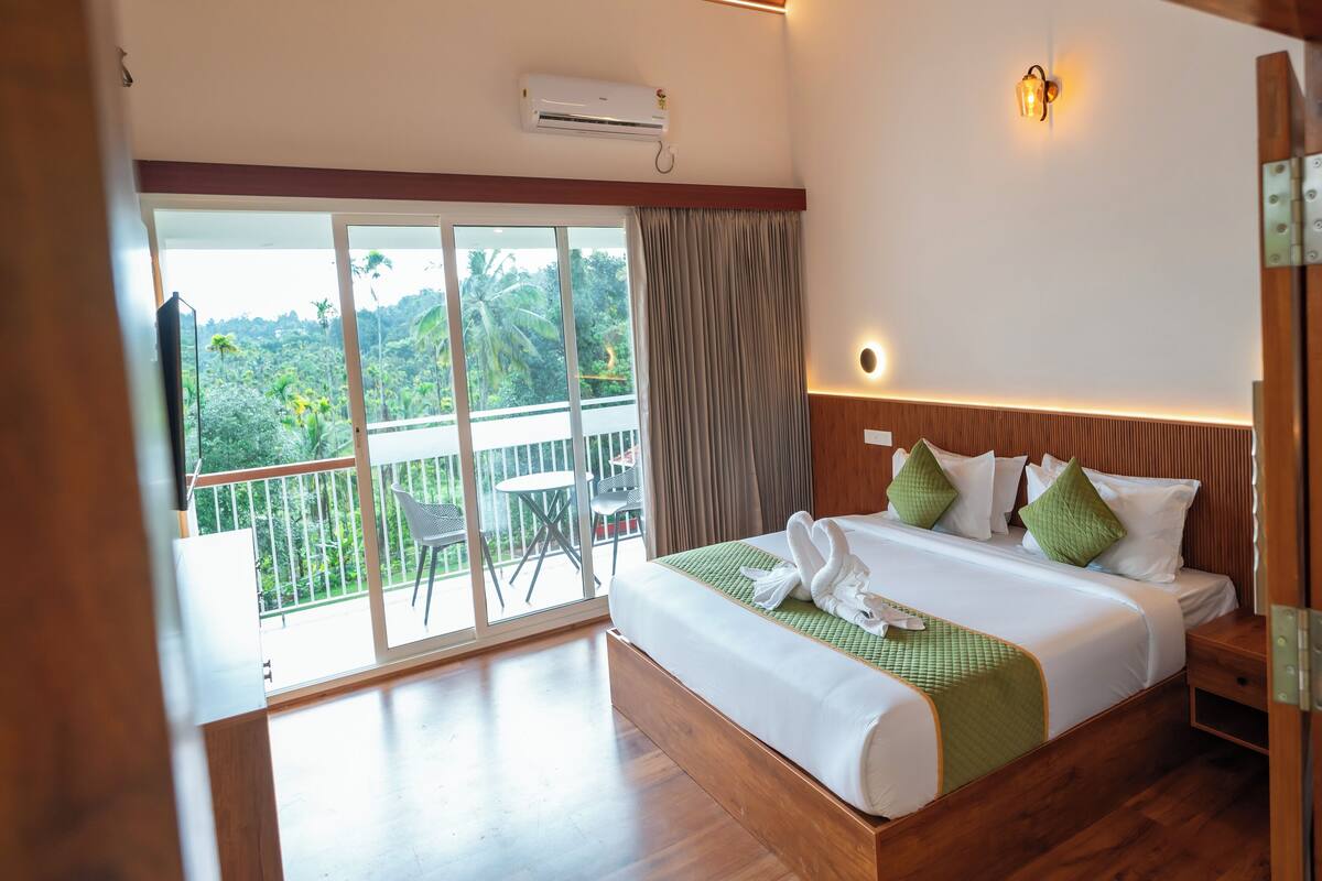 A hotel room with a large bed, a balcony with a view of the forest, and a ceiling fan.
