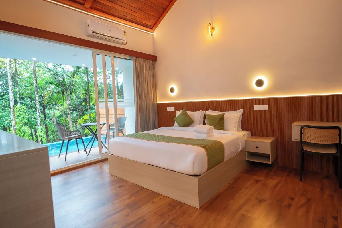 A modern hotel room with a large bed, a desk, and a view of a lush garden.