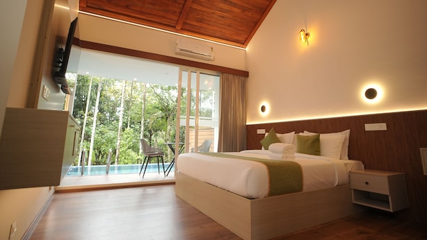 A modern hotel room with a large bed, a balcony with a view of greenery, and a wooden ceiling. - Theyila Wayanad (Sulthan Bathery)
