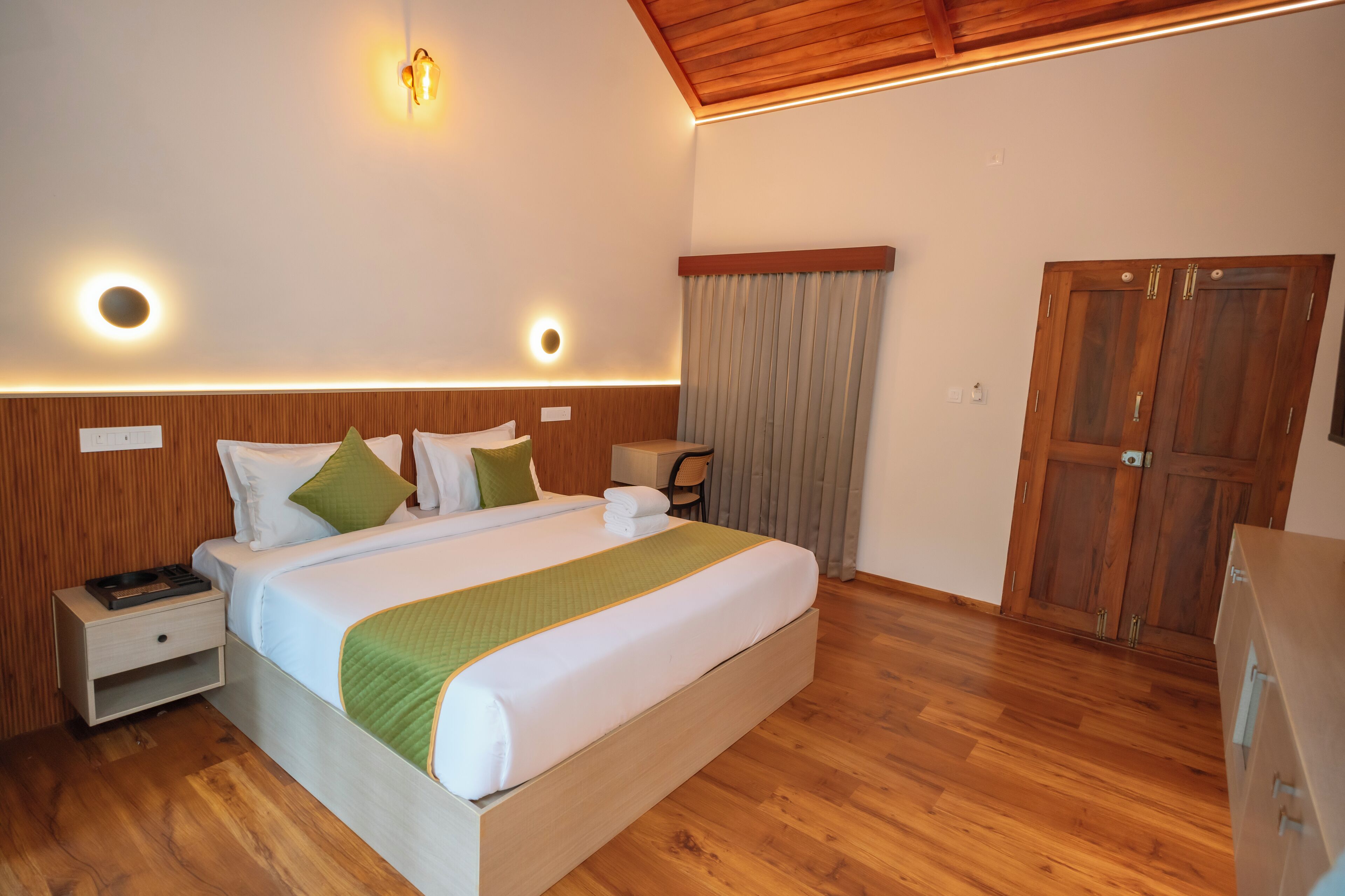 Een slaapkamer met een houten hoofdbord, een bed met wit beddengoed en groene accenten, een nachtkastje, een bureau, een kledingkast en een plafondlamp.