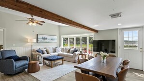 A spacious living room with a wooden ceiling, a large sofa set, a dining table with chairs, a television, and a view of the outdoors through large windows. - Honeoye Lake | Hot Tub | Kayak | Fire Pit (Honeoye)